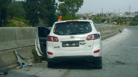 'Kami akan menyelamatkan siapa pun': Helm putih Suriah masuk setelah serangan mematikan 3 Mobil putih dengan lubang peluru duduk di penghalang pusat di jalan raya. Satu pintu terbuka, dan ada pakaian di tanah. Tidak ada orang yang terlihat dalam gambar