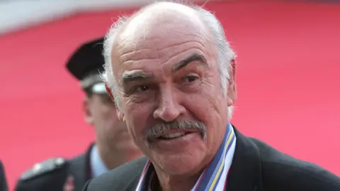 Getty Images Sean Connery di karpet merah pada tahun 2006. Dia memakai kumis dan mengenakan kemeja biru, putih dan kuning bergaris.