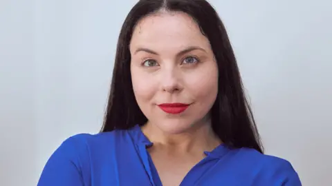 Amie Liebowitz Seorang wanita berambut hitam panjang tersenyum ke arah kamera. Dia berdiri di depan latar belakang putih dan mengenakan lipstik merah dan blus biru.