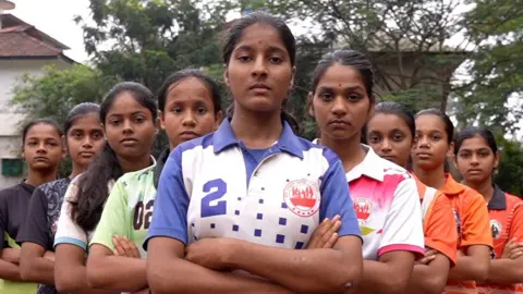 BBC sembilan gadis dari Kabaddi Club berdiri di Formasi V dengan tangan bersilang, melihat ke kamera. Meena ada di depan dengan empat gadis di setiap sisi mengipasi di belakangnya. Mereka mengenakan kemeja olahraga berwarna cerah.