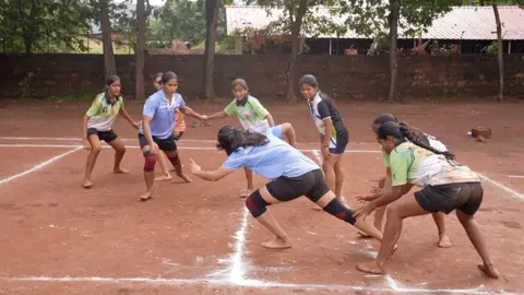 Tembakan aksi tujuh gadis bermain Kabaddi. Mereka mengenakan kemeja olahraga ungu, hijau dan hitam, celana pendek dan memiliki kaki telanjang. Pitchnya telanjang tanah dengan garis -garis putih yang ditandai di atasnya. 