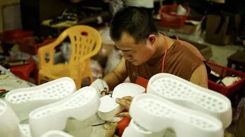 Xiqing Wang/ BBC Seorang pria bekerja di meja di pabrik kulit. Dia mengenakan kaos cokelat dan celemek merah, dengan sarung tangan putih tanpa jari di tangan kanannya. Dia dikelilingi oleh apa yang tampak seperti sol sepatu putih, salah satunya