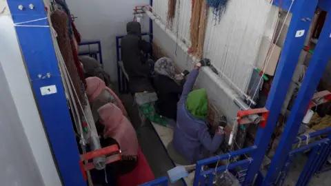 Dua baris tiga wanita muda, duduk kembali ke belakang, masing -masing menghadap alat tenun tinggi.