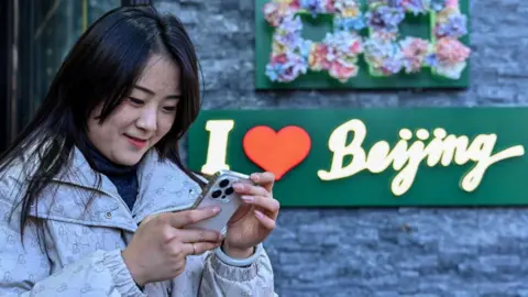 Getty Images Seorang wanita Cina memeriksa teleponnya, dengan tanda di latar belakang dibaca "Saya hati Beijing"