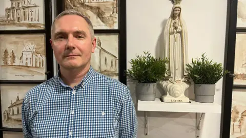 John Murphy Seorang pria berambut abu-abu pendek dengan kemeja kotak-kotak putih dan biru berdiri di depan serangkaian lukisan gereja, patung, dan beberapa pot tanaman. 