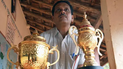 Daji Rajguru dengan kemeja yang diperiksa dengan dua piala emas besar di depannya. Trofi diembos dengan pola dan berbentuk seperti vas dengan tutup dan pegangan. 