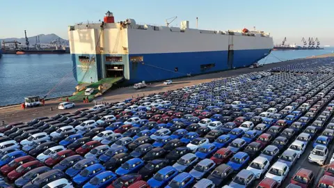 Getty Images Beberapa mobil berkendara ke perut kapal biru dan putih besar, yang merapat di sebelah tempat parkir yang penuh dengan mobil di pelabuhan Yantai pada 30 Maret 2025 di Yantai, Provinsi Shandong di Tiongkok.