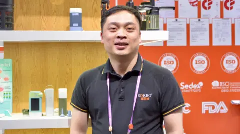 Xiqing Wang/ BBC Lionel Xu melihat langsung ke kamera, mengenakan T-shirt polo dengan logo perusahaannya di atasnya dan lanyard ungu di lehernya. Dia berdiri di depan elektronik di rak putih dengan latar belakang kayu