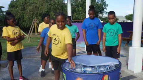 Gemma Handy Sekelompok anak-anak di T-shirt berwarna-warni bertuliskan jam tangan logo sekolah karena salah satu dari mereka memasukkan botol plastik ke tempat sampah daur ulang